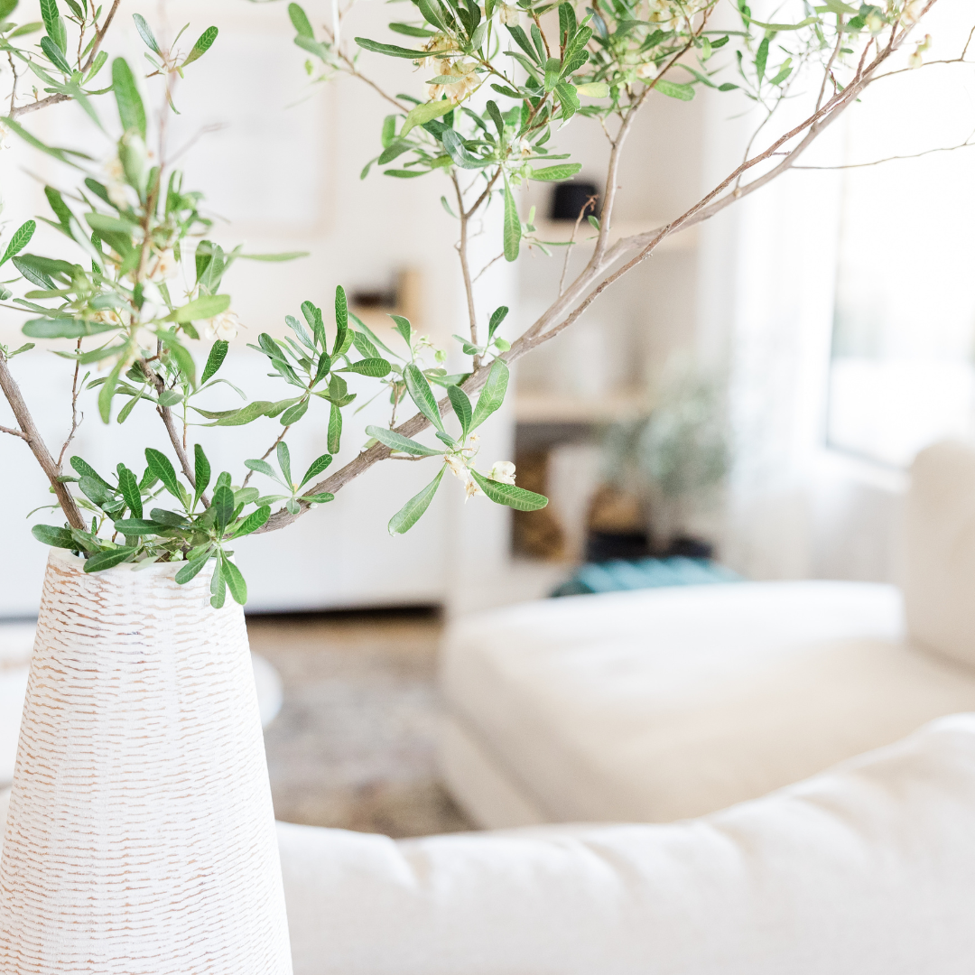 Ein weißer Vase mit grünen Zweigen steht im Vordergrund eines hellen Wohnzimmers mit einem Sofa im Hintergrund.